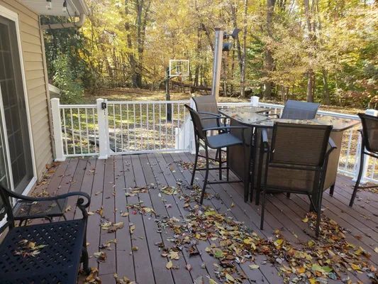 Deck with fallen leaves, a table with chairs, and a white railing in an outdoor setting with trees.