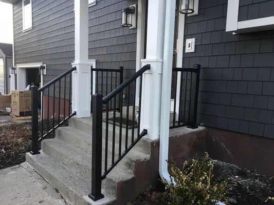 Exterior view of a house with concrete steps and black railing. Gray siding and white pillars are visible.