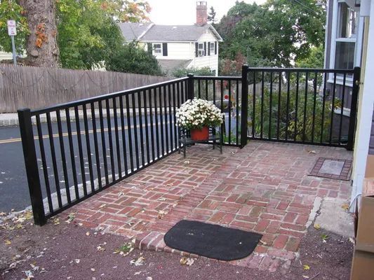 Black metal fence encloses a brick patio with a flower pot. Street and house in the background.