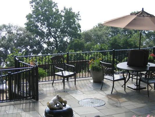 Patio with wrought-iron furniture, umbrella, and a view of trees through a black railing.