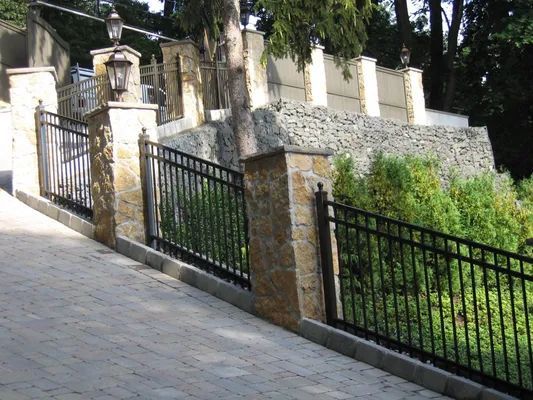 Black metal fence on stone pillars along a sloped brick road.