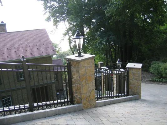 Fence with stone columns, decorative lamps, and wrought iron railings bordering a paved driveway near a house.