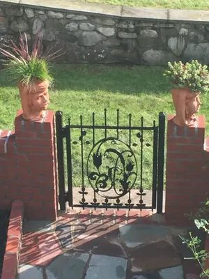 Black wrought iron garden gate between brick pillars, topped with decorative planters with greenery.
