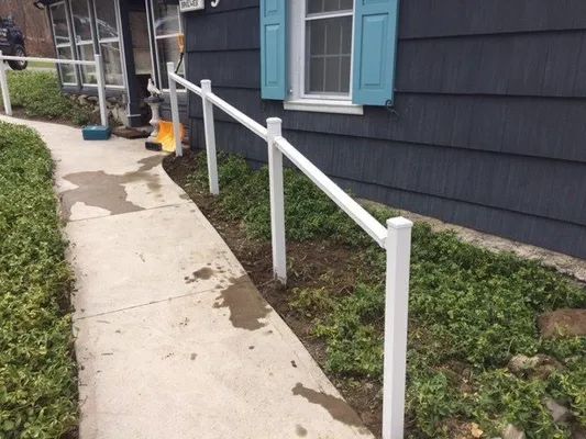 White handrail alongside a concrete sidewalk leading to a blue-sided building with a window and shutters.