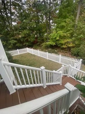 White picket fence encloses a grassy area. Deck with white railings and brown flooring leads down. Trees in background.