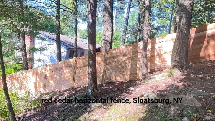 Red cedar horizontal fence in Sloatsburg, NY, partially obscures a house amidst trees in a wooded setting.