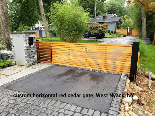 Horizontal red cedar gate on a driveway in West Nyack, NY, with a stone pillar, black posts, and greenery.