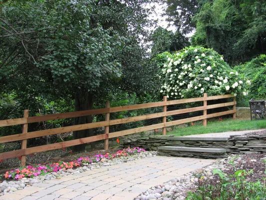 Wooden split-rail fence alongside a brick pathway and flower bed, leading to steps, with trees in background.