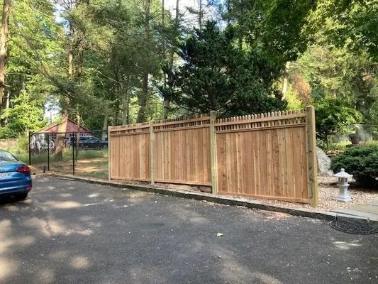 Wooden privacy fence in a parking area, partially obscuring a backyard with trees.