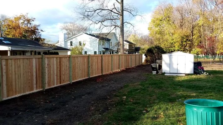 Wooden fence bordering a gravel path and grassy yard, with houses and trees in the background.