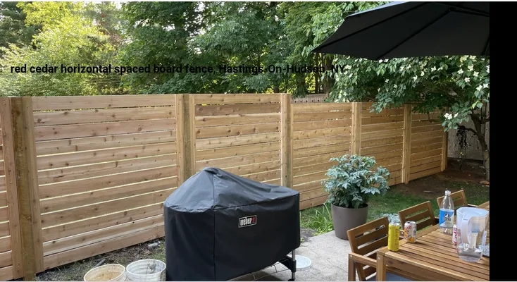 Horizontal red cedar fence encloses a patio with a grill, table, potted plant, and umbrella.