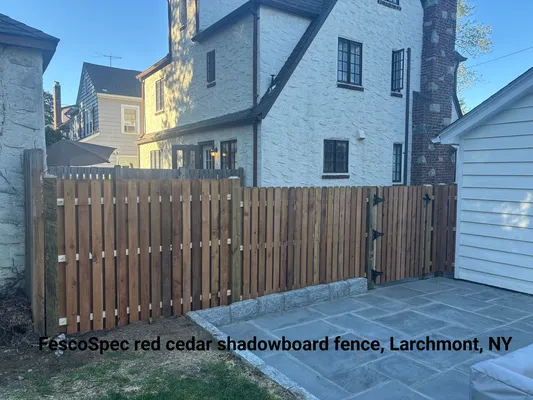 Red cedar shadowboard fence in Larchmont, NY, framing a backyard patio with stone pavers and a large house.