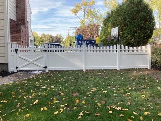 White picket fence with gate, bordering a grassy yard next to a brick building.