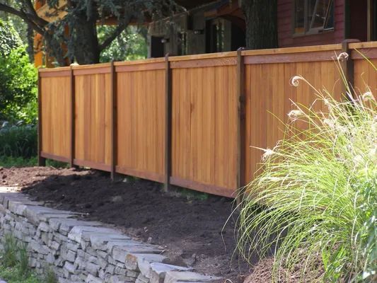 Wooden fence with dark brown posts sits atop a stone retaining wall. Brown soil and green plants are in foreground.