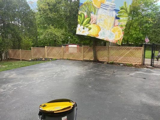 Lattice fence surrounds a paved driveway. A lemonade-themed mural hangs in the background. A trash can is in the foreground.