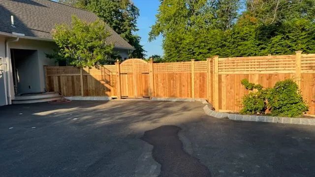 Wooden fence surrounding a driveway. A gate and house are visible. Trees are in the background.