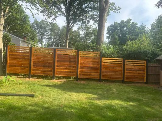 Wooden horizontal slat fence in a grassy yard, surrounded by trees under a cloudy sky.