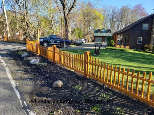 Red cedar picket fence along a road in Blauvelt, NY, with a blue pickup truck parked behind it on a sunny day.