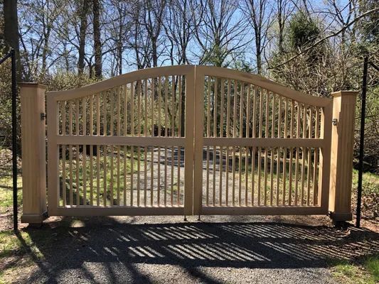 Wooden double gate, arched top, with vertical spindles, set in a wooded area on a sunny day.