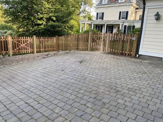 Cobblestone driveway with picket fence, leading to a light-colored house.