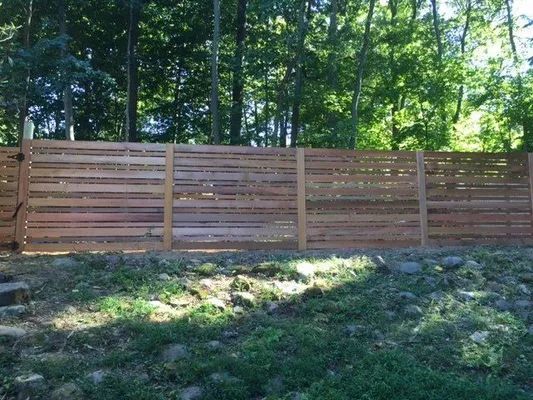 Wooden horizontal slat fence in front of a forest with green grass and foliage.