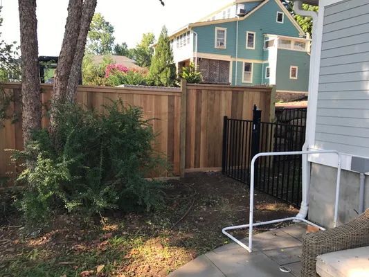 Wooden fence with black metal gate next to a light-colored house. A white handrail is present.