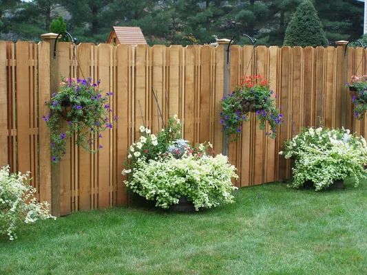 Wooden fence with hanging flower baskets and planted flowers in a grassy yard.