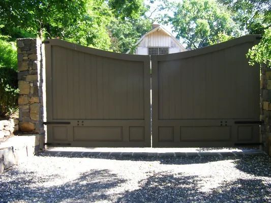 Large, beige wooden driveway gates set between stone pillars; a house is visible in the background.