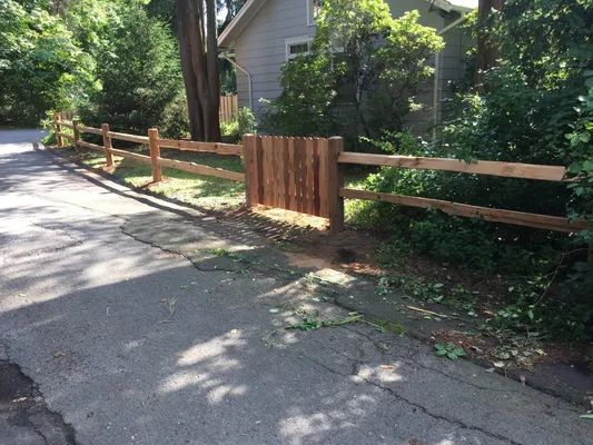 Wooden split-rail fence with a vertical plank gate alongside a paved road, greenery in the background.