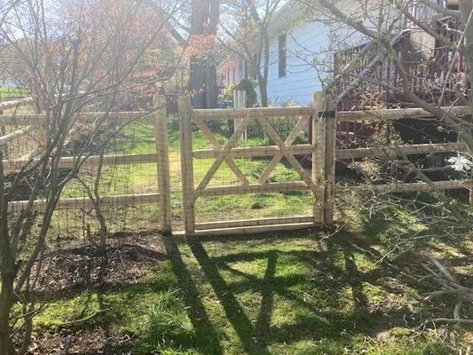 Wooden gate in a split-rail fence; green grass and trees in the background, sunny day.