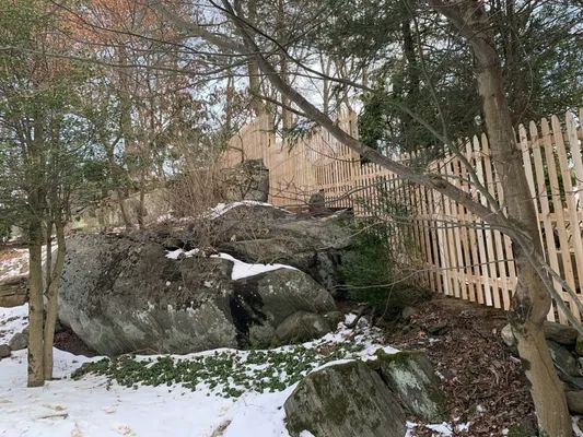 Large rock formation next to a wooden fence and bare trees in a snow-covered yard.