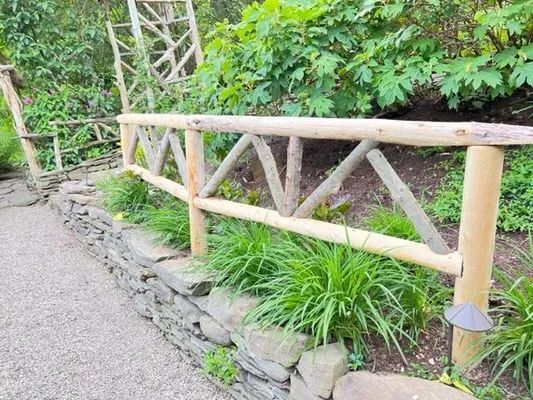 Rustic wooden fence in a garden, built with logs and angled supports, next to a stone wall and path.