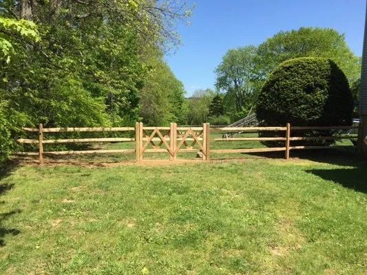 Wooden split-rail fence with gate in a grassy yard, trees in background, sunny day.