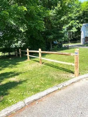 Wooden split-rail fence alongside a road in a grassy yard, with trees in the background.
