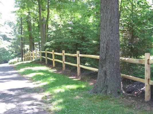 Wooden split-rail fence alongside a paved path and trees in a wooded area.