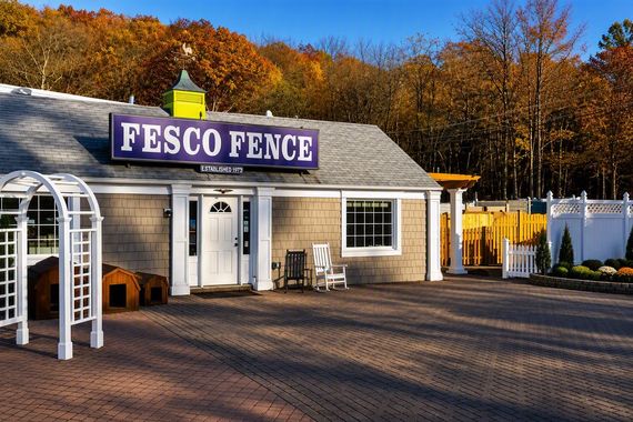FESCO Fence building with sign, blue door, and small display of outdoor furniture. Background of trees and blue sky.