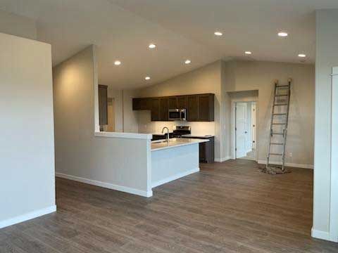 Interior view of a modern kitchen and living area. Brown cabinets, white island, wood-look floors, and recessed lighting.