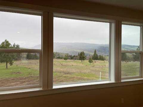 Three white-framed windows offer a view of a green field, trees, and distant mountains under a cloudy sky.