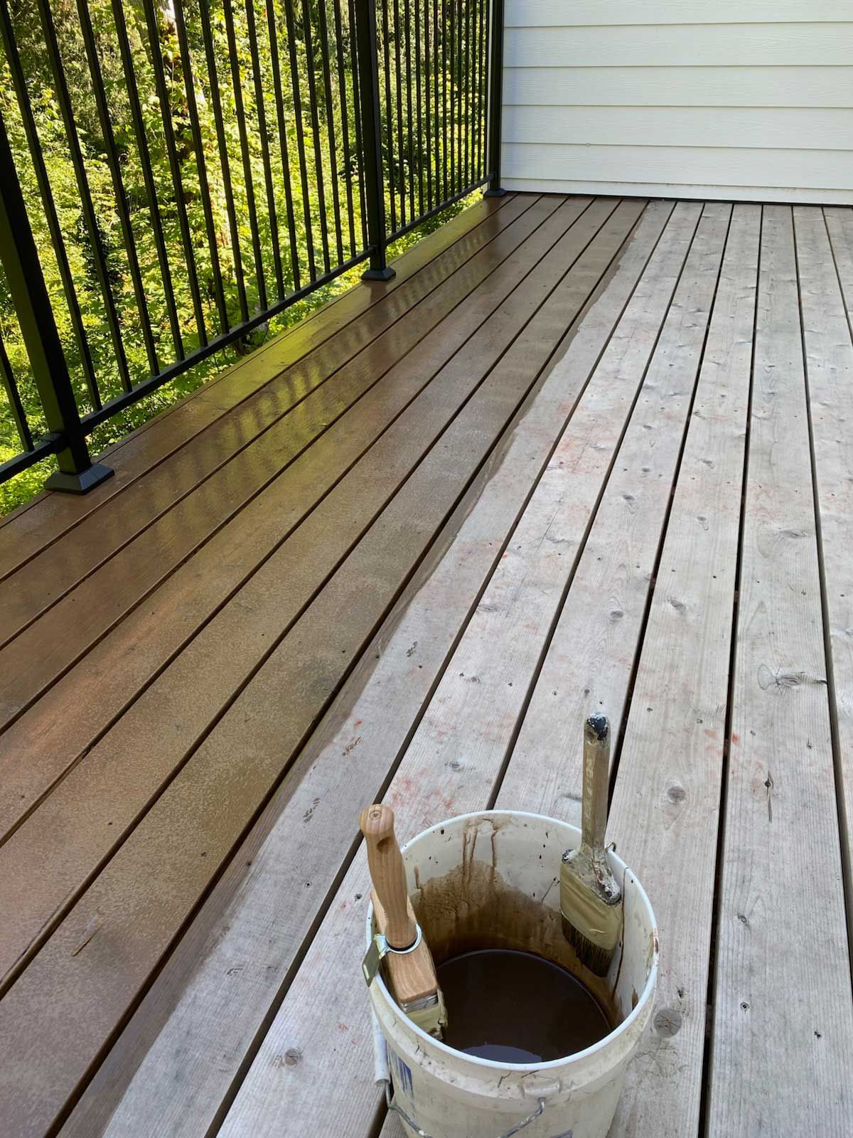 A wooden deck being stained brown with a paint bucket and brushes. Black railing and green foliage in the background.