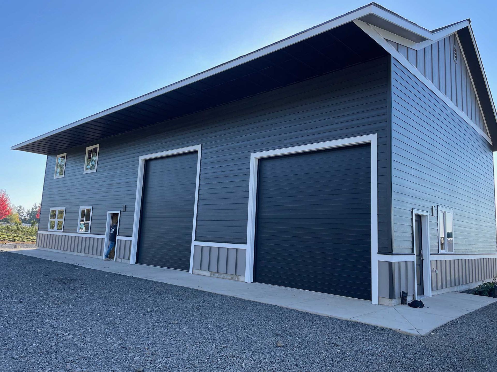 A large gray barn with black garage doors, a person stands on the side in gravel under a blue sky.