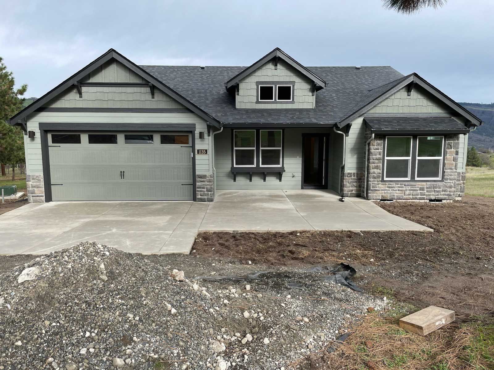 Green ranch-style house with gray roof and garage door, stone accents, and unfinished yard.