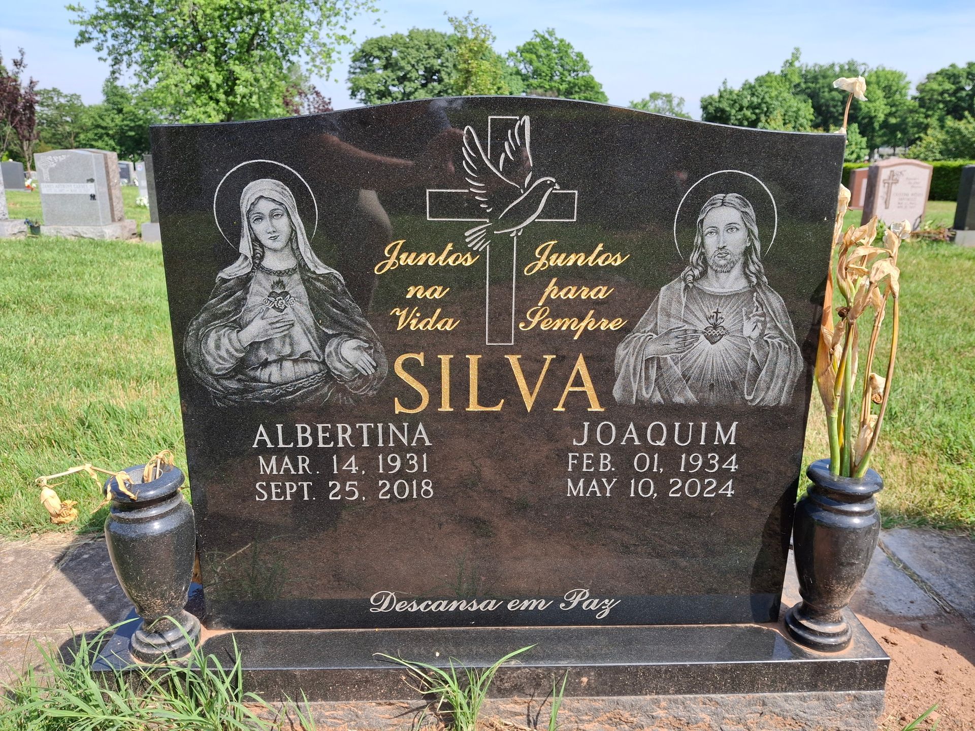 Black granite memorial headstone with engraved names and crosses in a cemetery, flanked by small vases