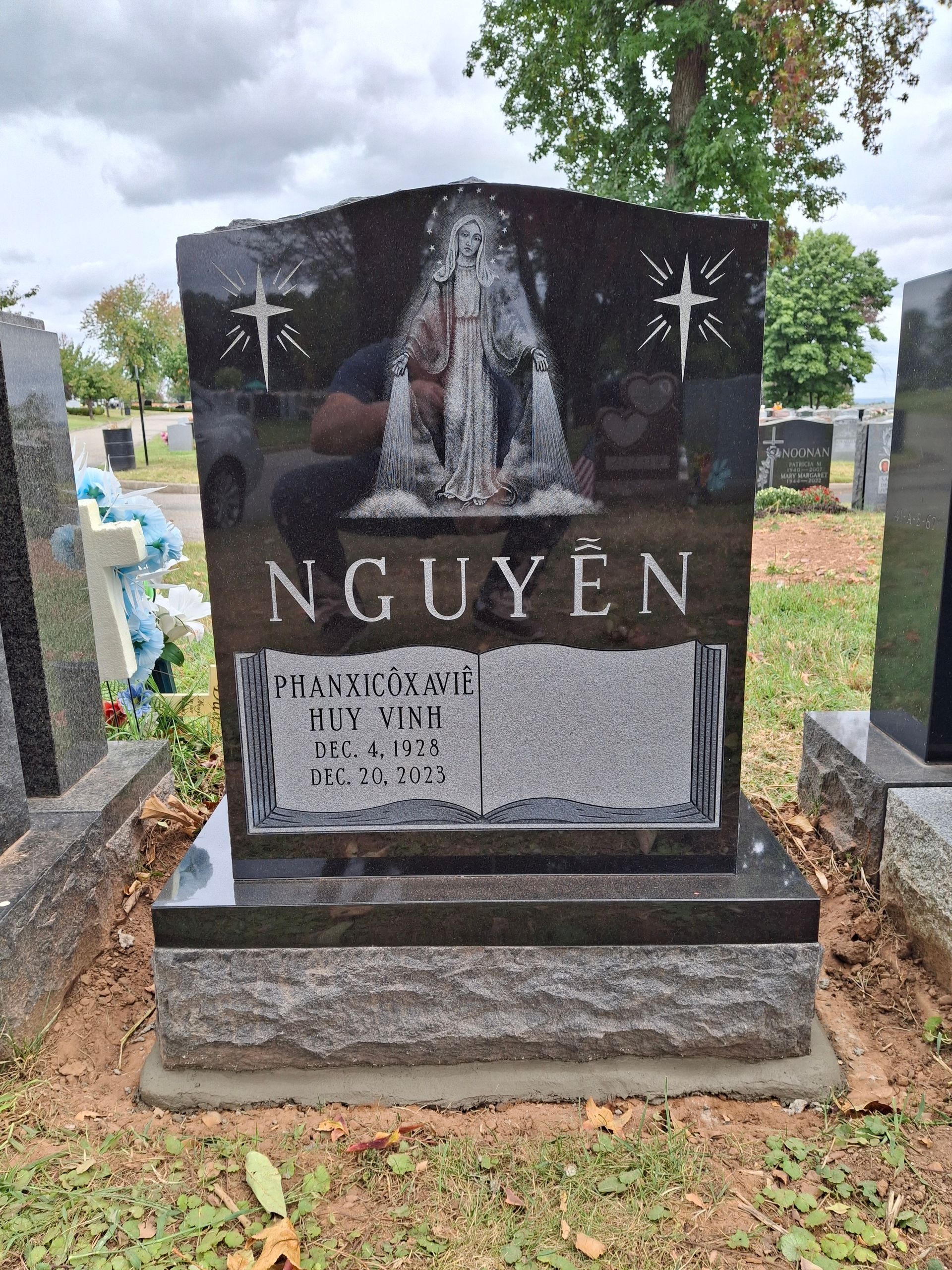 Black granite gravestone with “NGUYEN,” centered cross design, in a cemetery with trees and nearby markers.