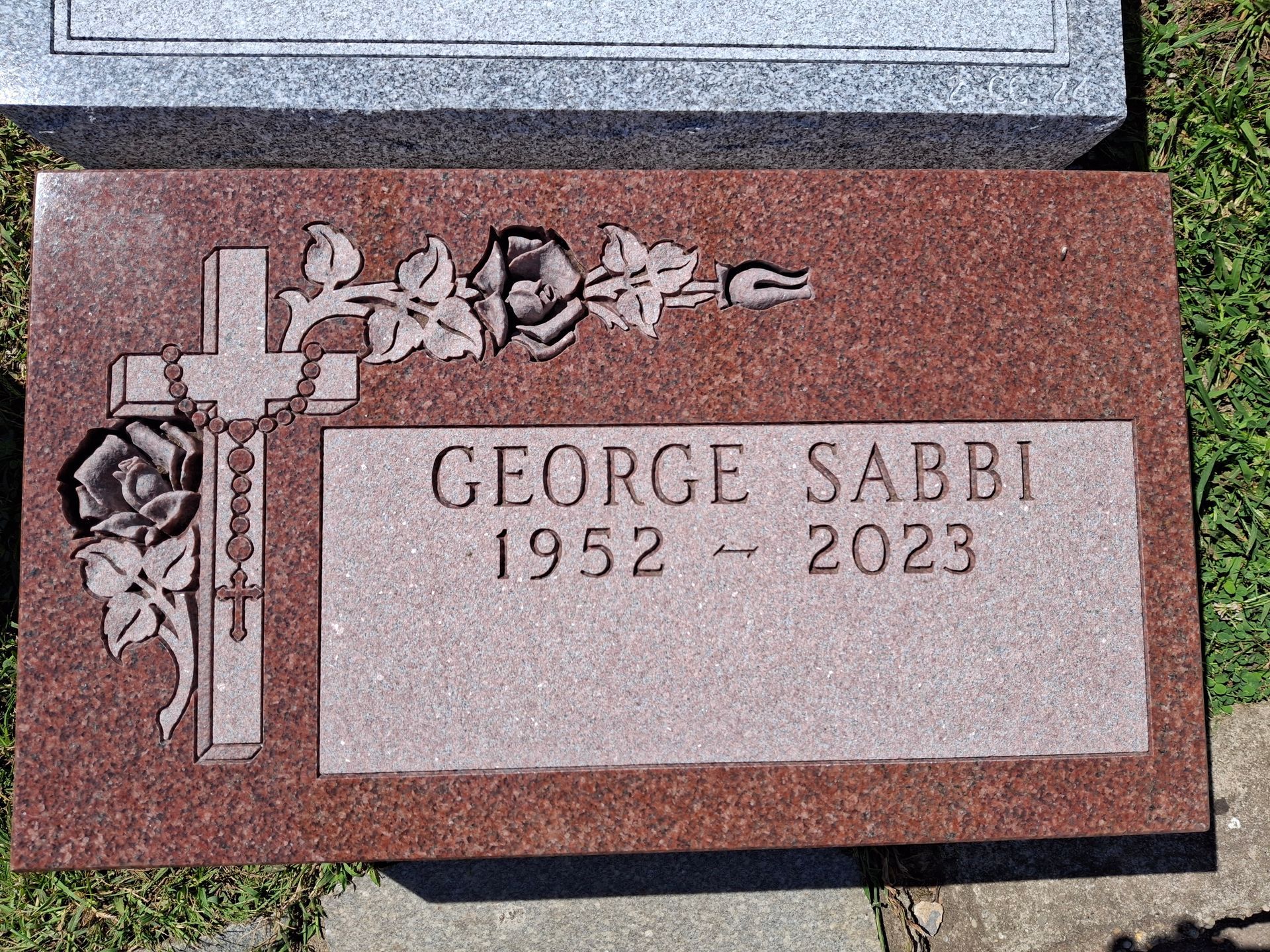 Red granite memorial plaque with a cross, roses, and the inscription “GEORGE SABI 1952–2023”