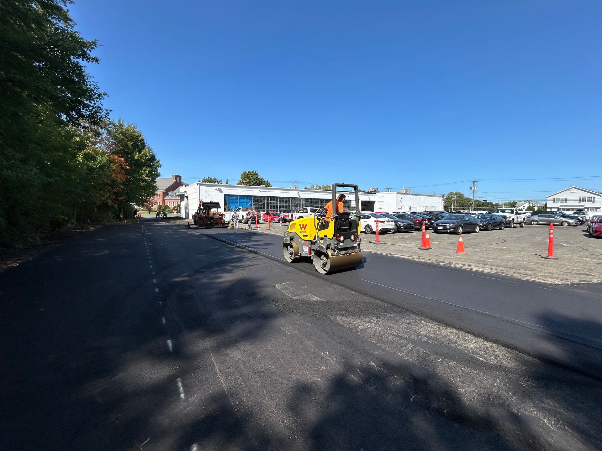 A yellow construction roller paving a fresh asphalt surface in a parking lot on a sunny day.