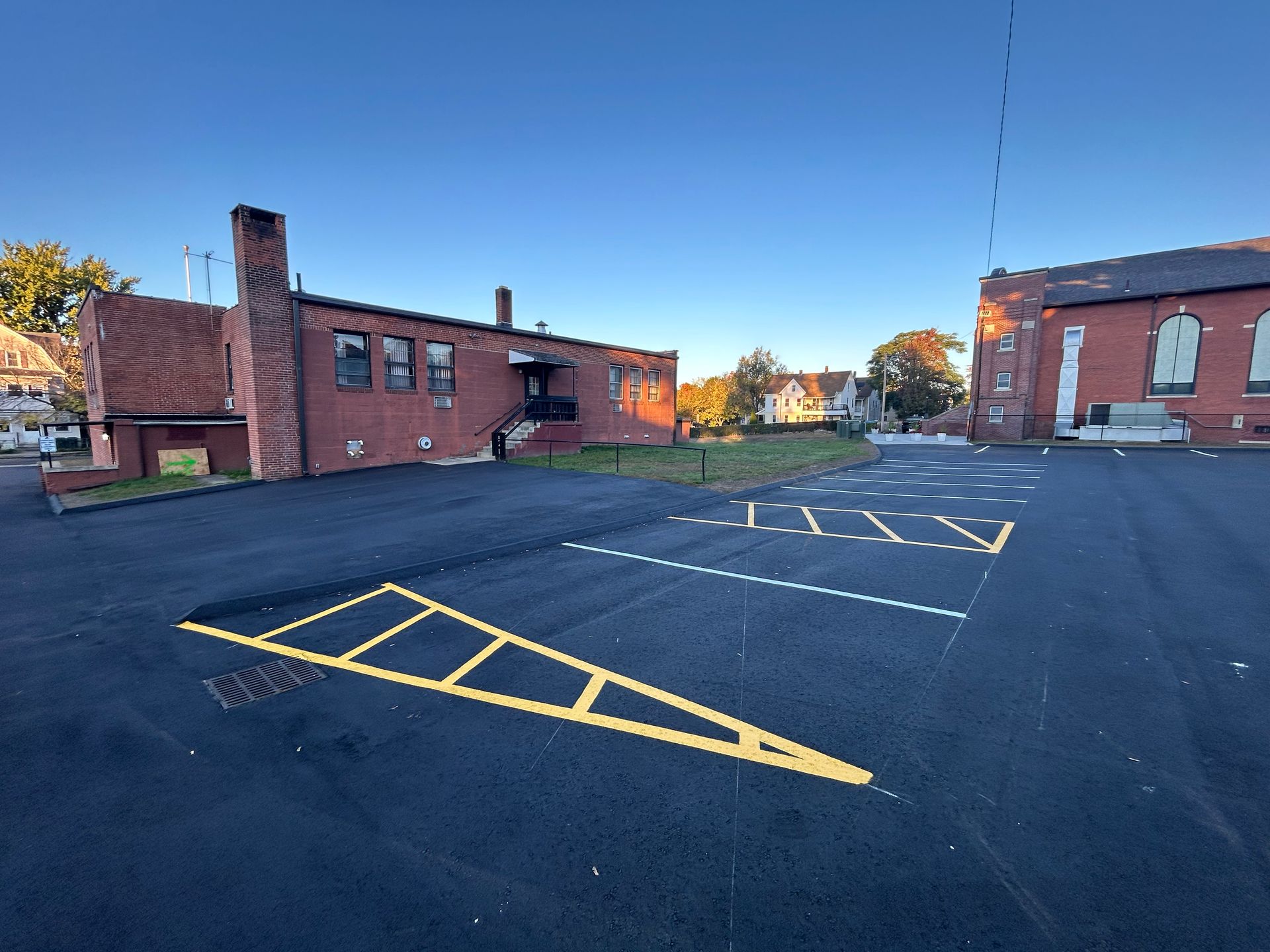 A newly paved asphalt parking lot with yellow-painted safety markings between two red brick buildings on a sunny day.