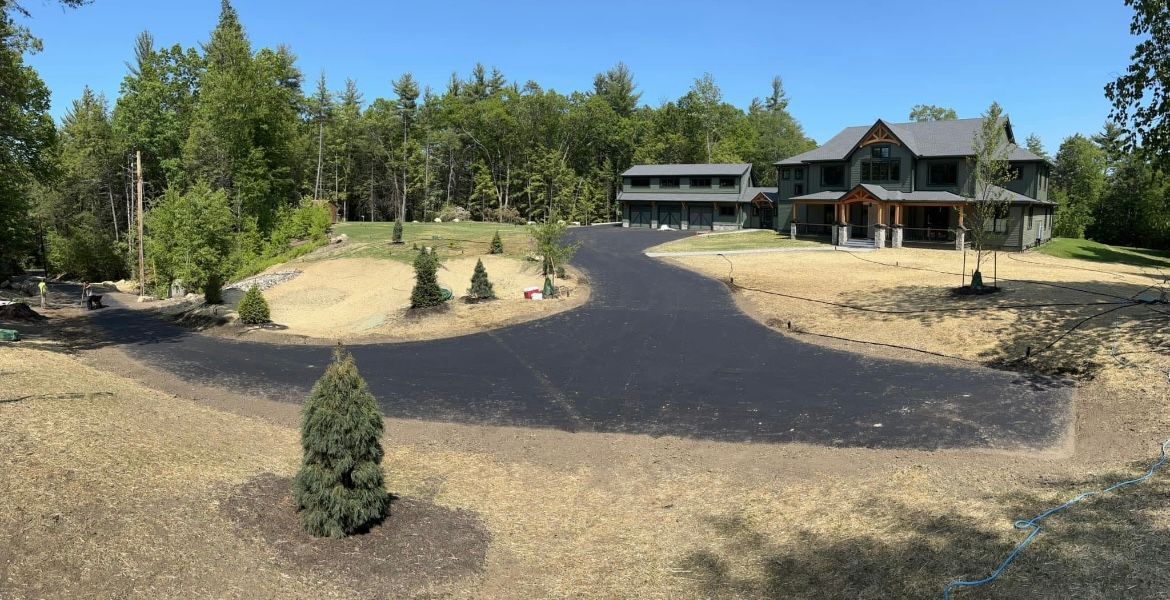 A freshly paved, wide black asphalt driveway leads toward a two-story home and detached garage surrounded by trees.