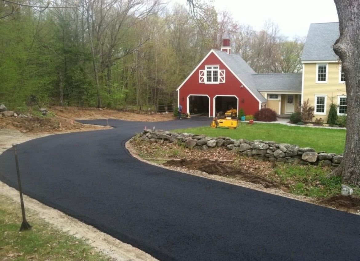 A smooth, newly paved black asphalt driveway leads toward a red barn-style garage next to a yellow house.