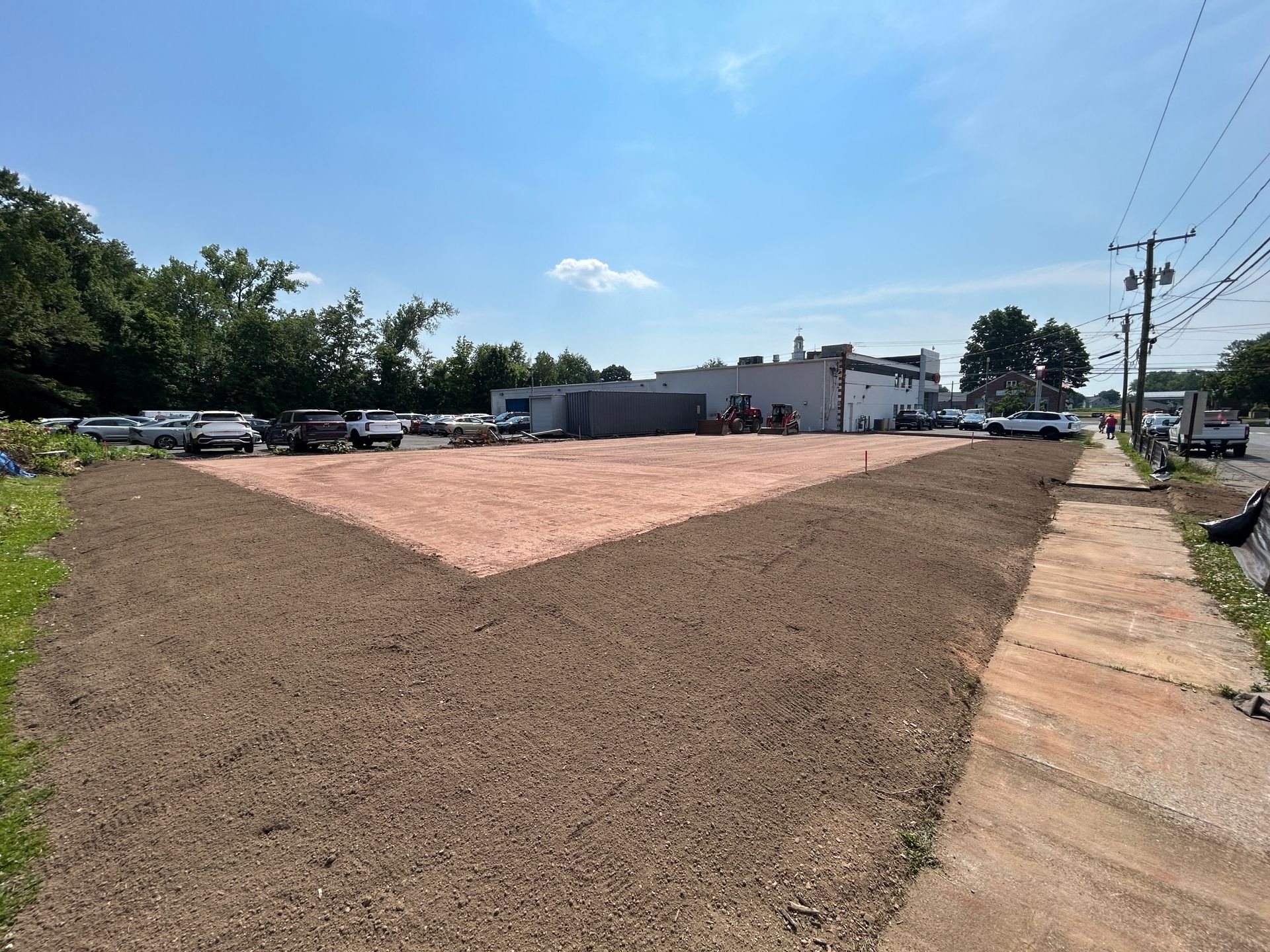 A vacant lot with a pink gravel center surrounded by brown soil, bordered by a sidewalk on the right and a building beyond.