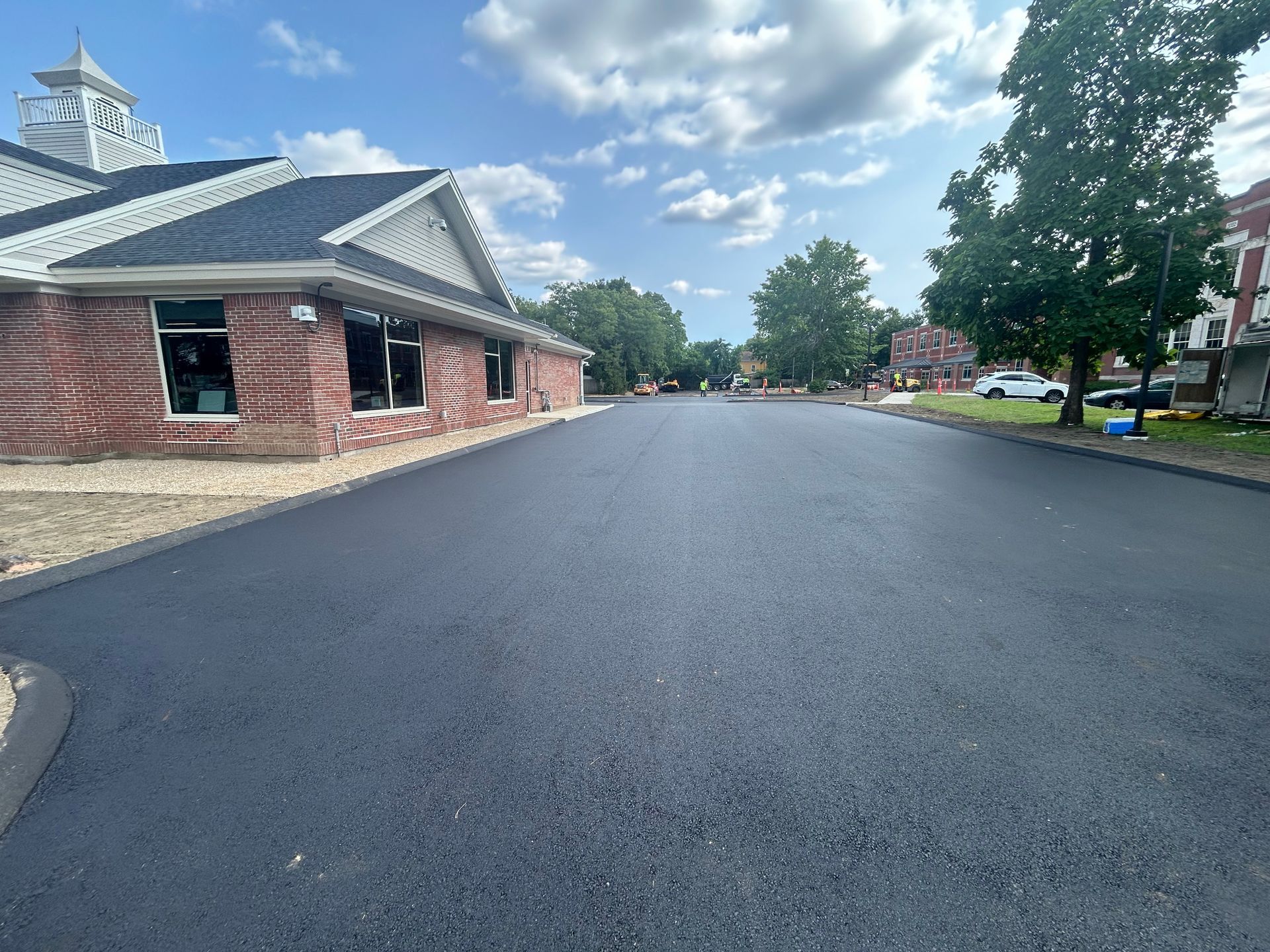 A freshly paved asphalt road runs past a brick building under a sunny, blue sky with clouds.